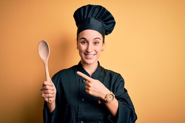 Young beautiful chef woman wearing cooker uniform and hat holding wooden spoon very happy pointing...