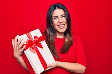 Young beautiful brunette woman holding birthday gift over isolated red background looking positive and happy standing and smiling with a confident smile showing teeth