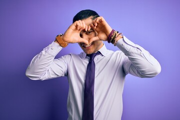 Young brazilian businessman wearing elegant tie standing over isolated purple background Doing heart shape with hand and fingers smiling looking through sign