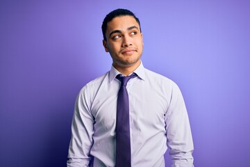 Young brazilian businessman wearing elegant tie standing over isolated purple background smiling looking to the side and staring away thinking.