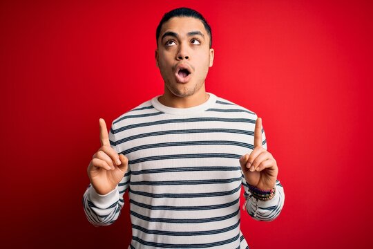 Young brazilian man wearing casual striped t-shirt standing over isolated red background amazed and surprised looking up and pointing with fingers and raised arms.