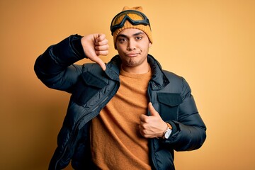 Young brazilian skier man wearing snow sportswear and ski goggles over yellow background Doing thumbs up and down, disagreement and agreement expression. Crazy conflict