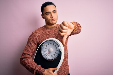 Young brazilian man doing diet to lose weigth holding scale over isolated pink background with...