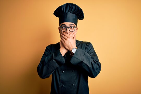 Young brazilian chef man wearing cooker uniform and hat over isolated yellow background shocked covering mouth with hands for mistake. Secret concept.