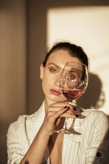 Sensual young woman in white jacket holding glass of wine near her face