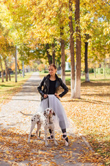 Fototapeta premium Ballerina with Dalmatian dog in the Park. Woman ballerina in a white ballet skirt and black leather jacket dancing in pointe shoes in autumn park with her spotty dalmatian dog.