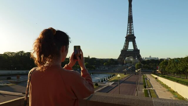 Young woman taking vertical pictures of the eiffel tower with her phone during early morning in summer, arc shot from behind