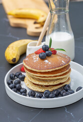 Pancake with fresh berries on white plate for breakfast with banana and milk on black table