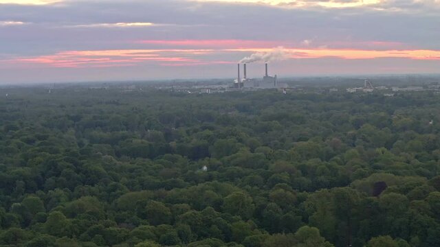 Drone shot backwards of a power plant at the wide horizon over green trees of Munichs Englischer Garten including a lake.