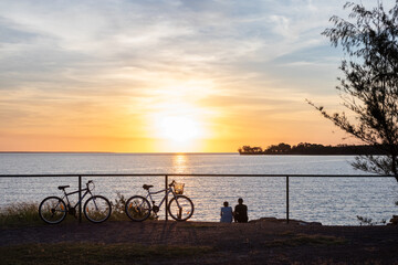 Darwin, Northern Territory, Australia; May 2020: Couple watching sunset. Bicycles on the side. First day after covid-19 restrictions are partially relaxed in Darwin, Northern Territory NT, Australia - obrazy, fototapety, plakaty