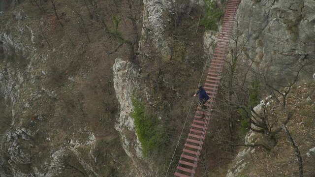 Man crossing rope bridge between rock cliffs in Turda Gorge while hiking the Via Ferrata. Bridge shakes and wobbles as man walks from plank to plank over deep valley.