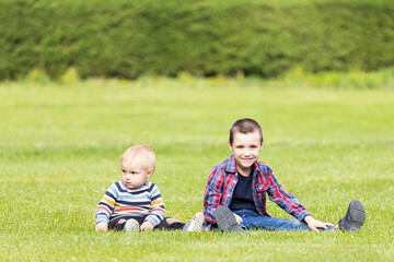 Two cheerful boy brothers of different ages play fun, sit on the grass on a warm summer day. The concept of friendship of brothers and happy childhood.