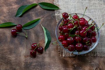 red cherries in a transparent glass bowl on a wooden background, with green leaves of a cherry tree. Summer berries. Yummy. © Yuliia
