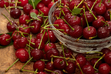 red cherries in a transparent glass bowl on a wooden background, with green leaves of a cherry tree. Summer berries. Yummy.