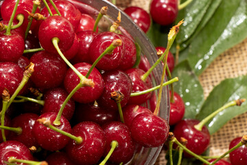 red cherries in a transparent glass bowl on a wooden background, with green leaves of a cherry tree. Summer berries. Yummy.