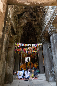 Cambodia; February 2020: Statue of stone buddha with shoulder covered by yellow fabrics. Colorful flags hanging from ceiling, buddhist people pray on the floor. Banteay Kdei temple, Angkor, Siem Reap