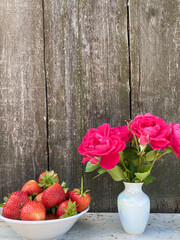 Bowl with ripe strawberries and a decorative vase with bright flowers on a background of abstract wooden wall. Free space.