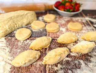 Uncooked dumplings with strawberries and dough on an abstract wooden surface. The process of preparing a traditional meal of flour dough.