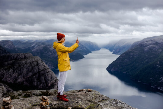Alone blonde woman hiker in yellow raincoat stays on the top of the Preikestolen mountain (Preacher's Pulpit or Pulpit Rock) with a lot of tourists and shoots photos by phone on Lysefjord background
