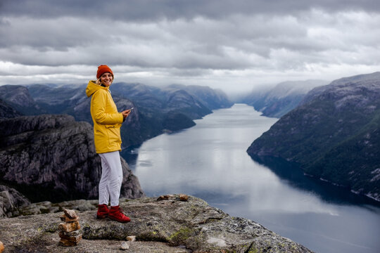 Smiling blonde woman hiker in yellow raincoat stays on the top of the Preikestolen mountain (Preacher's Pulpit or Pulpit Rock) with a lot of tourists and shoots photos by phone on Lysefjord background
