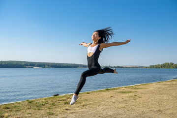 Fototapeta premium Smiling young female gymnast is jumping in split outdoors near the lake. Healthy lifestyle