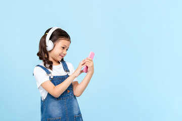 Cute 8 year-old girl wearing headphones listening to music online from mobile phone isolated on light blue background