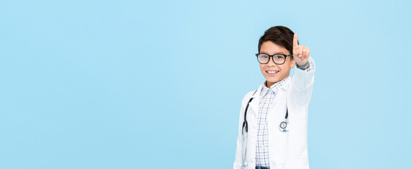 Little kid doctor smiling and pointing hand upward isolated on light blue background