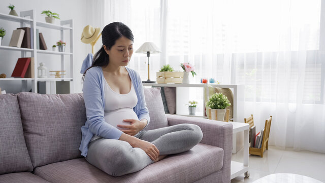 Pretty pregnant woman resting on sofa alone at home in day time. frowning maternity lady sit on couch and feel depression at apartment. future motherhood holding big belly and look pensive thoughtful
