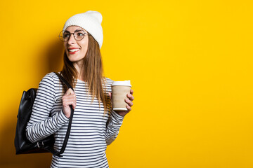 Portrait of a happy young woman in glasses and hat, holds a paper cup with a coffee on a yellow background