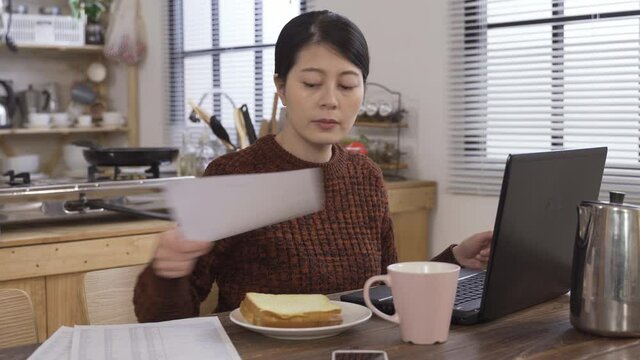 asian millennial woman is filing tax with laptop at breakfast. taiwanese female sitting at dining table is entering information on withholding certificate to find out how much tax she should pay.