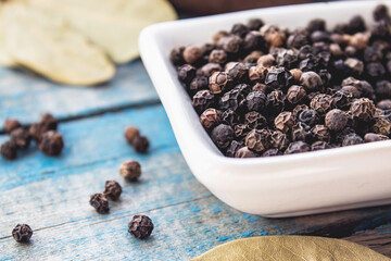 Black pepper in a bowl near a bay leaf on a background of wooden blue boards.