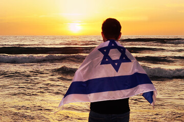 Concept: Mediterranean Sea in Israel at sunset. Opposite the sea, out of focus, is a young Israeli patriot with the flag of Israel on his shoulders