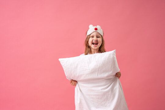 Cheerful Little Girl In A Sleep Mask Looks Out From Behind A Pillow