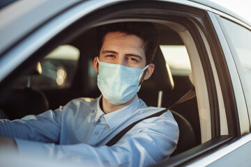 Fototapeta na wymiar Young boy taxi driver gives passenger a ride wearing sterile medical mask. A man in the car behind the steering wheel works during coronavirus pandemic. Social distance and health safety concept.