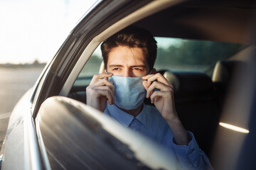 Young businessman takes a taxi and looks out of the car window adjusting sterile medical mask. Man sits on the back seat of taxi and takes a ride during coronavirus pandemic. Social distance concept.