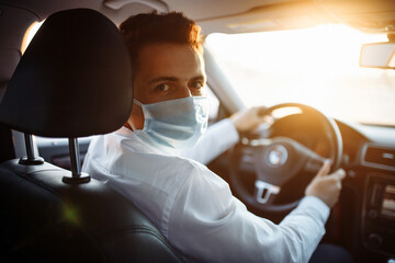 Young man taxi driver steers the car during coronavirus pandemic wearing sterile medical mask. A boy drives a car through the traffic on the sunset. Social distance and health care concept.