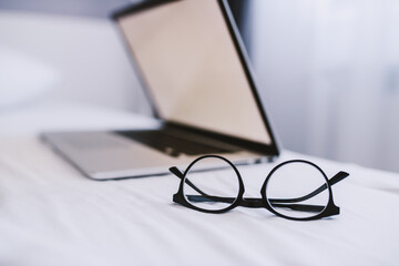 Picture of eyeglasses and laptop on bed in bedroom. Selective focus on eyeglasses. - obrazy, fototapety, plakaty