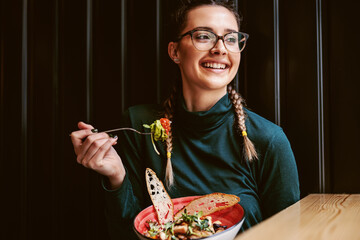 Young attractive teenage girl with healthy habits sitting in restaurant, holding bowl with salad...