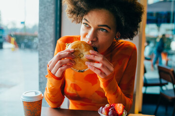 Young beautiful afro woman indoors restaurant biting hamburger - hungry diverse woman sitting...