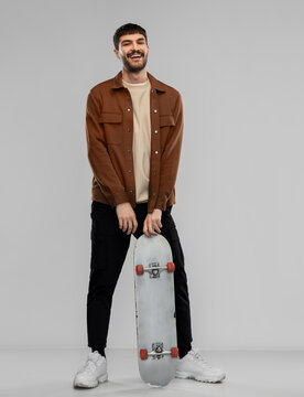 Leisure And People Concept - Happy Smiling Young Man In Brown Jacket And Black Pants With Skateboard Over Grey Background