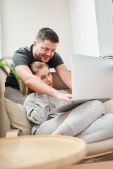 Fototapeta premium Young blonde sitting on beige armchair with laptop and brunet man standing near her in apartment