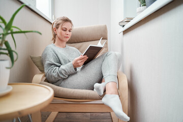 Young girl reading book while sitting on beige armchair in apartment