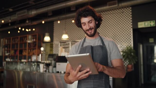 Young successful cafe owner standing in cafe wearing black striped apron using digital tablet while accepting online order