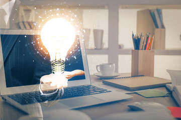 Computer on desktop in office with bulb icon hologram. Multi exposure. Concept of idea.