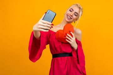 Naklejka premium European blond girl in a red dress takes a selfie on the phone and holds a red heart out of paper on an orange studio background