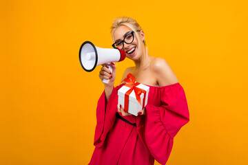 attractive blonde girl in a red dress shouts the news in a loudspeaker holding a gift box with a red ribbon on a yellow background