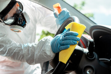 specialist in hazmat suit and respirator cleaning car interior with antiseptic spray and rag during covid-19 pandemic