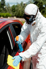 specialist in hazmat suit cleaning car with antiseptic spray and rag during covid-19 pandemic