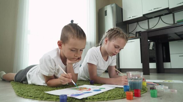 Children at home, a boy and a girl sit on the floor and draw on paper, drawing with watercolor paint.