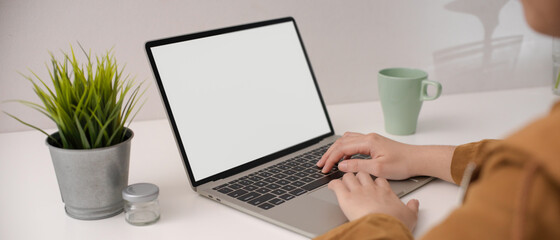 Female employee typing on blank screen laptop on white simple worktable with mug and decorations